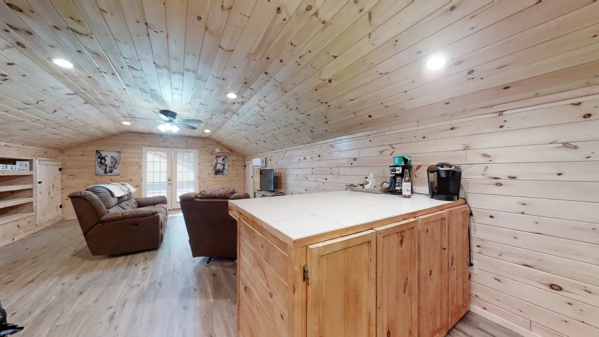 A comfortable seating area with a kitchenette, clad in natural wood paneling, creating a cozy, inviting space for rest and socializing. Large windows suggest a connection to the surrounding rural environment.