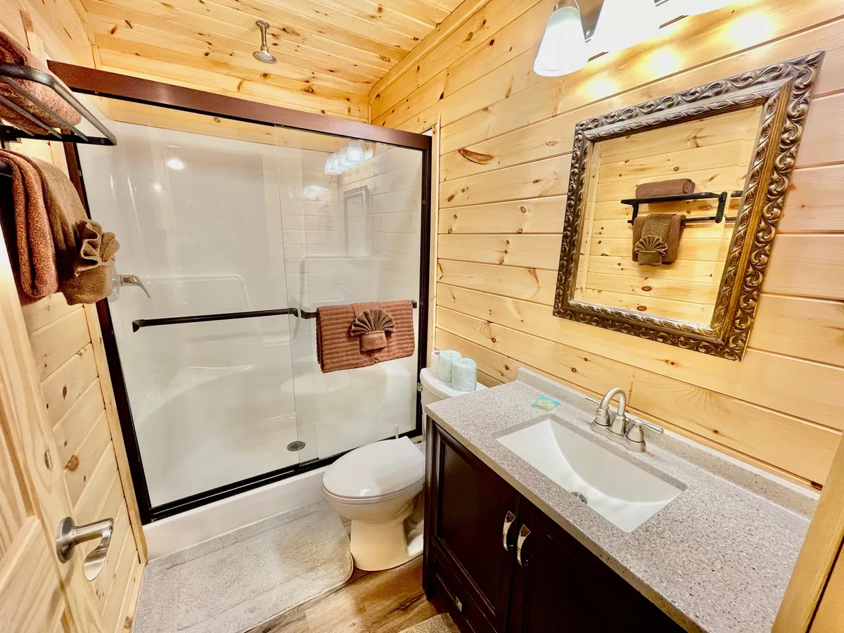 A bright ensuite bathroom featuring a glass shower enclosure, a framed mirror over the vanity with a modern sink, wooden walls, and neatly arranged towels. The design is both functional and inviting, creating a welcoming space for personal routines.