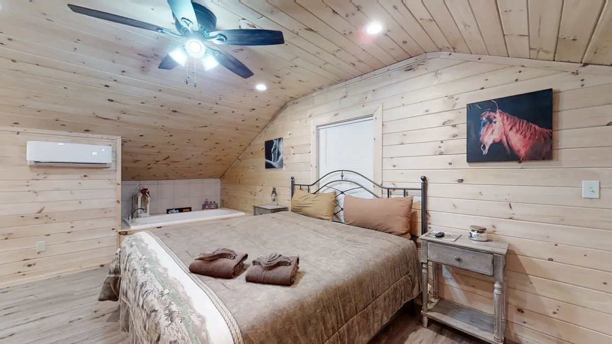 A cozy bedroom featuring rustic wooden walls, a comfortable bed, a ceiling fan, and a prominent painting of a horse that adds equestrian flair. The atmosphere is inviting and warm, ideal for relaxation after a day outdoors.
