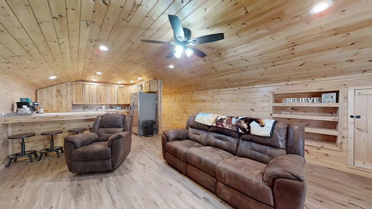 This image captures the heart of the living room, featuring a spacious brown sofa and a matching comfortable recliner set against a rustic wooden backdrop. The 'BELIEVE' wall decor and partial view of the kitchen area enhance the feeling of a cozy, lived-in space. Soft lighting and a warm color palette reinforce the inviting, rustic atmosphere.