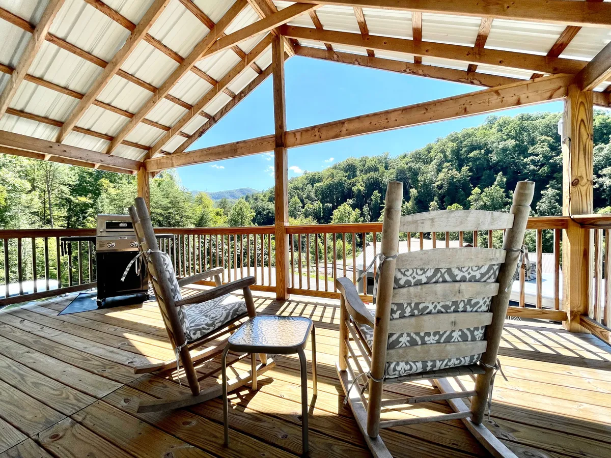 A private deck showcasing multiple rocking chairs and a grill, set against a backdrop of rolling hills and mountain views. The scene evokes restful outdoor living, with possibilities for barbecuing or stargazing.