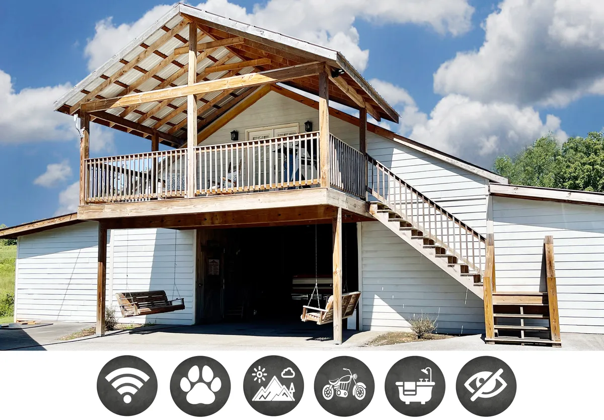The image showcases the exterior of a cozy barndominium apartment set against the backdrop of rolling hills. A spacious covered porch and balcony are the central focal points, featuring comfortable swing chairs perfect for relaxing. The sturdy staircase and inviting roof structure add rustic charm, while the setup suggests opportunities for enjoying the mountain air, sipping coffee, and sharing outdoor meals.