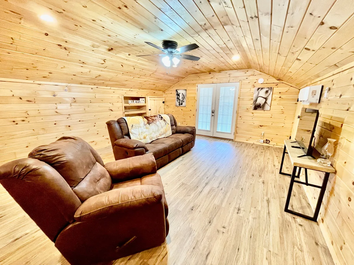 The image shows a reclining sofa positioned in a living room with wood-paneled walls, a TV stand with flat-screen TV, decorative wall art, and double doors that let in natural light. The setup suggests a versatile space for watching TV, reading, or enjoying a peaceful moment.