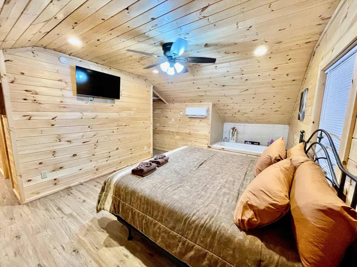 This image showcases a plush bed with brown bedding and pillows, a cozy wooden-walled and ceiling interior, a ceiling fan above the bed, a TV mounted on the wall, and a small kitchenette area to the side. The overall ambiance is warm and inviting, ideal for relaxation and intimate retreats.