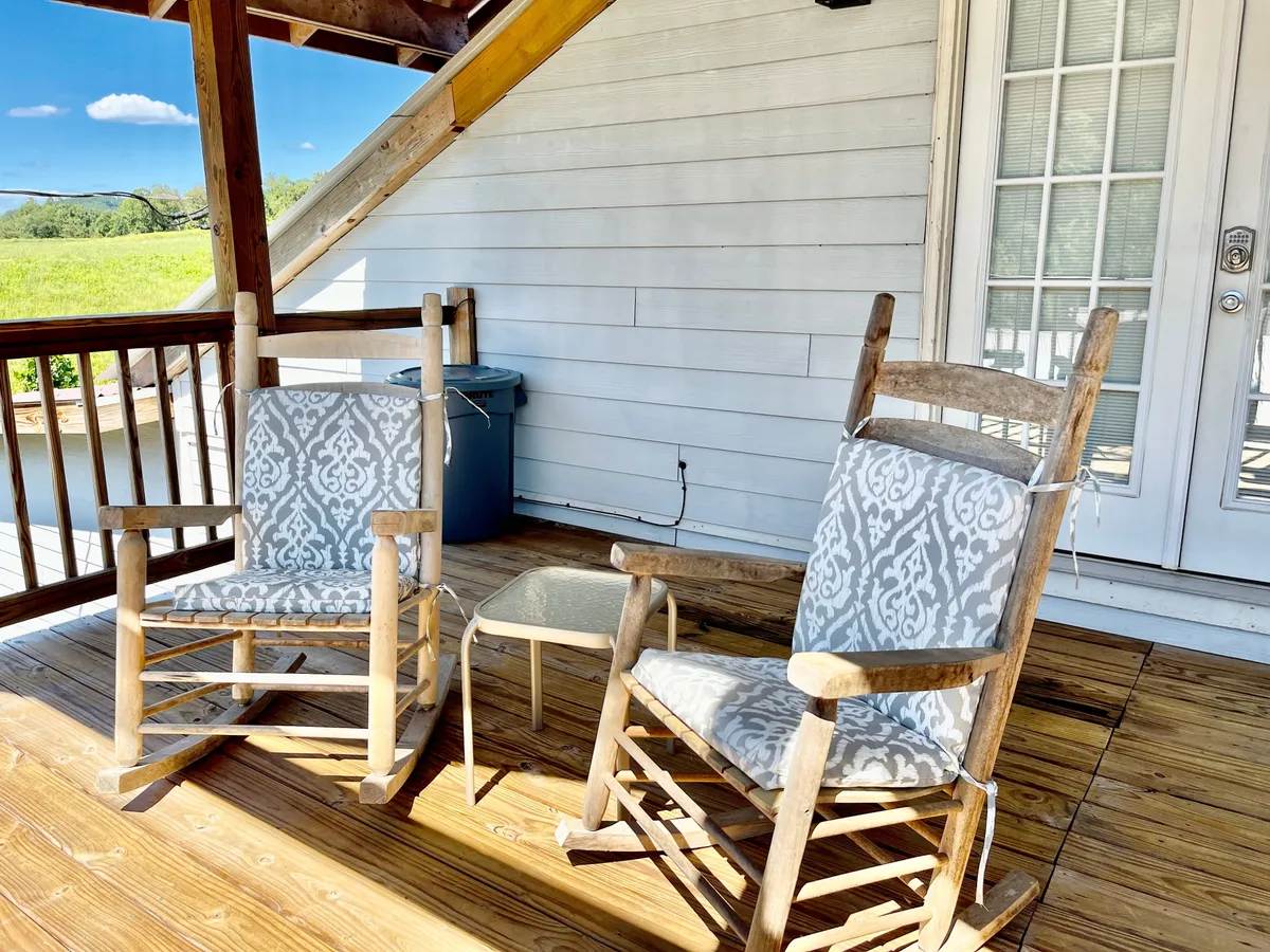 A cozy front porch featuring classic wooden rocking chairs and a small table, set against a rustic wooden deck and entryway. The porch overlooks serene outdoor space, inviting relaxation and connection with nature.